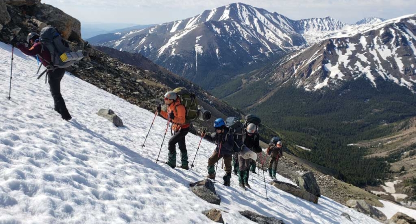 A group of students wearing helmets and carrying backpacks use trekking poles as they navigate over a snowy incline.